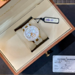 White and gold Ulysse Nardin wristwatch in an open beige presentation box with a hanging tag attached and a warranty card nearby.