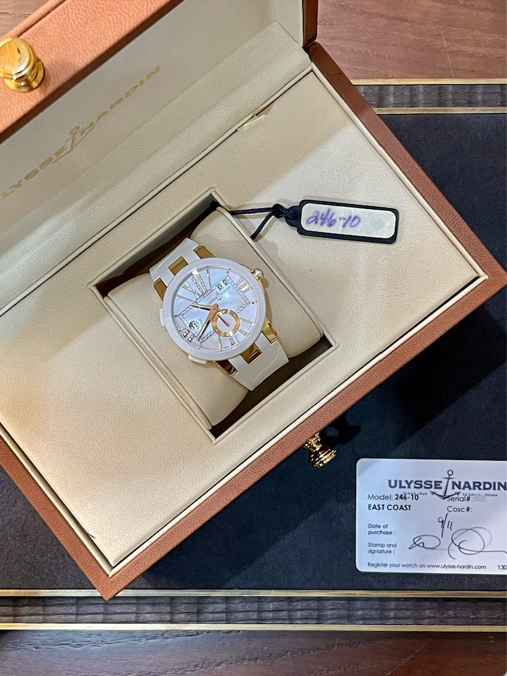 White and gold Ulysse Nardin wristwatch in an open beige presentation box with a hanging tag attached and a warranty card nearby.