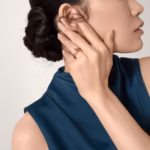 Woman with a sleek bun wearing rose-gold rings on her fingers, touching her ear in a close-up portrait with a blue sleeveless top.