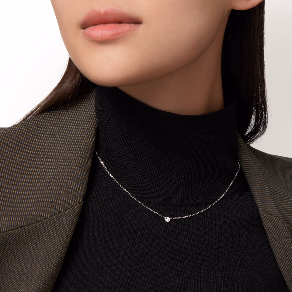 Close-up of a woman wearing a delicate silver necklace with a small bead pendant over a black turtleneck and brown pinstripe blazer.