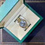 Open green jewelry box showing a two-tone gold-and-silver wristwatch with Roman numerals inside the beige interior