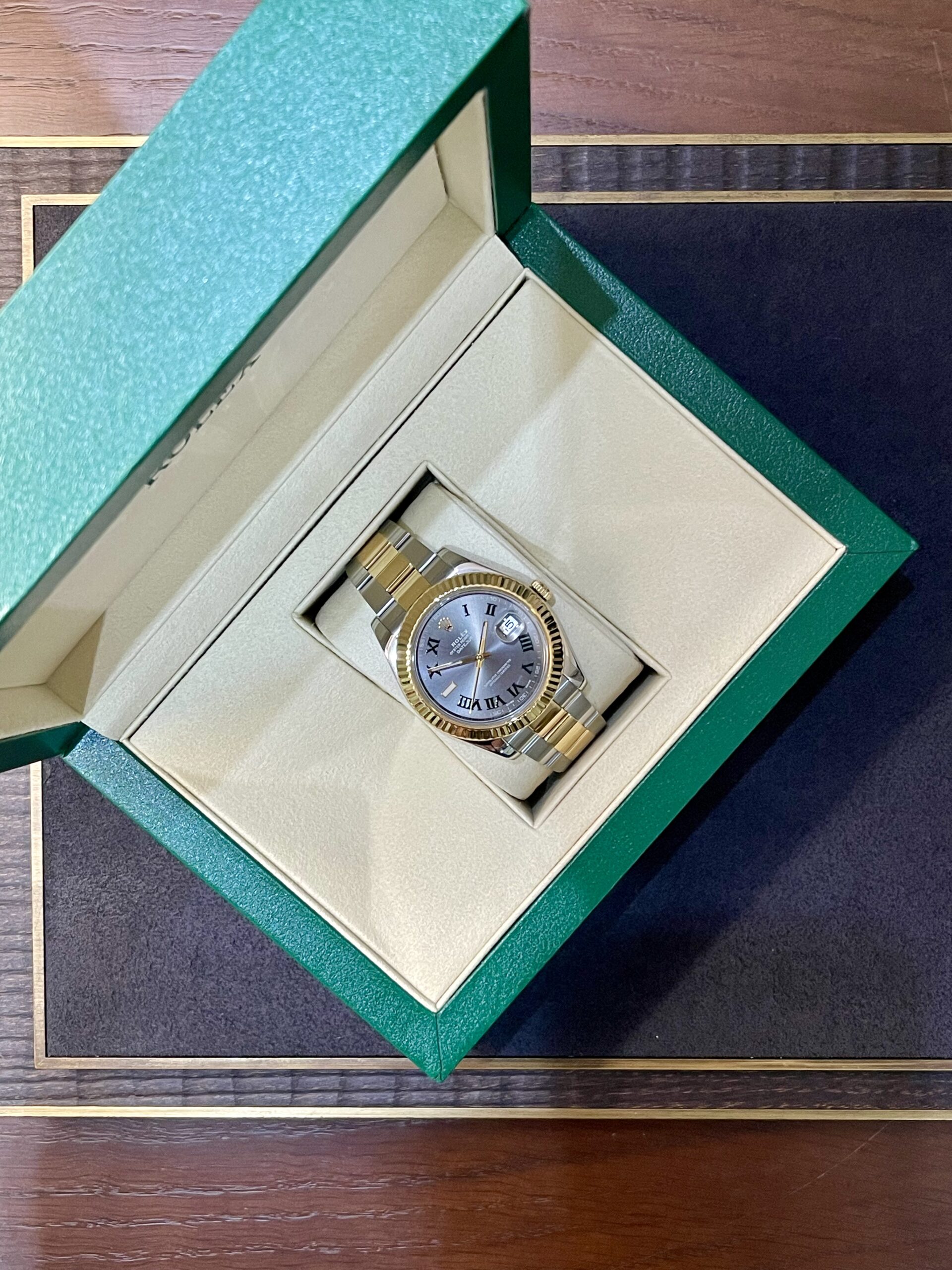 Open green jewelry box showing a two-tone gold-and-silver wristwatch with Roman numerals inside the beige interior