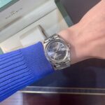 Silver Rolex Datejust on a wrist with a blue sweater sleeve; a Rolex box is visible in the background.