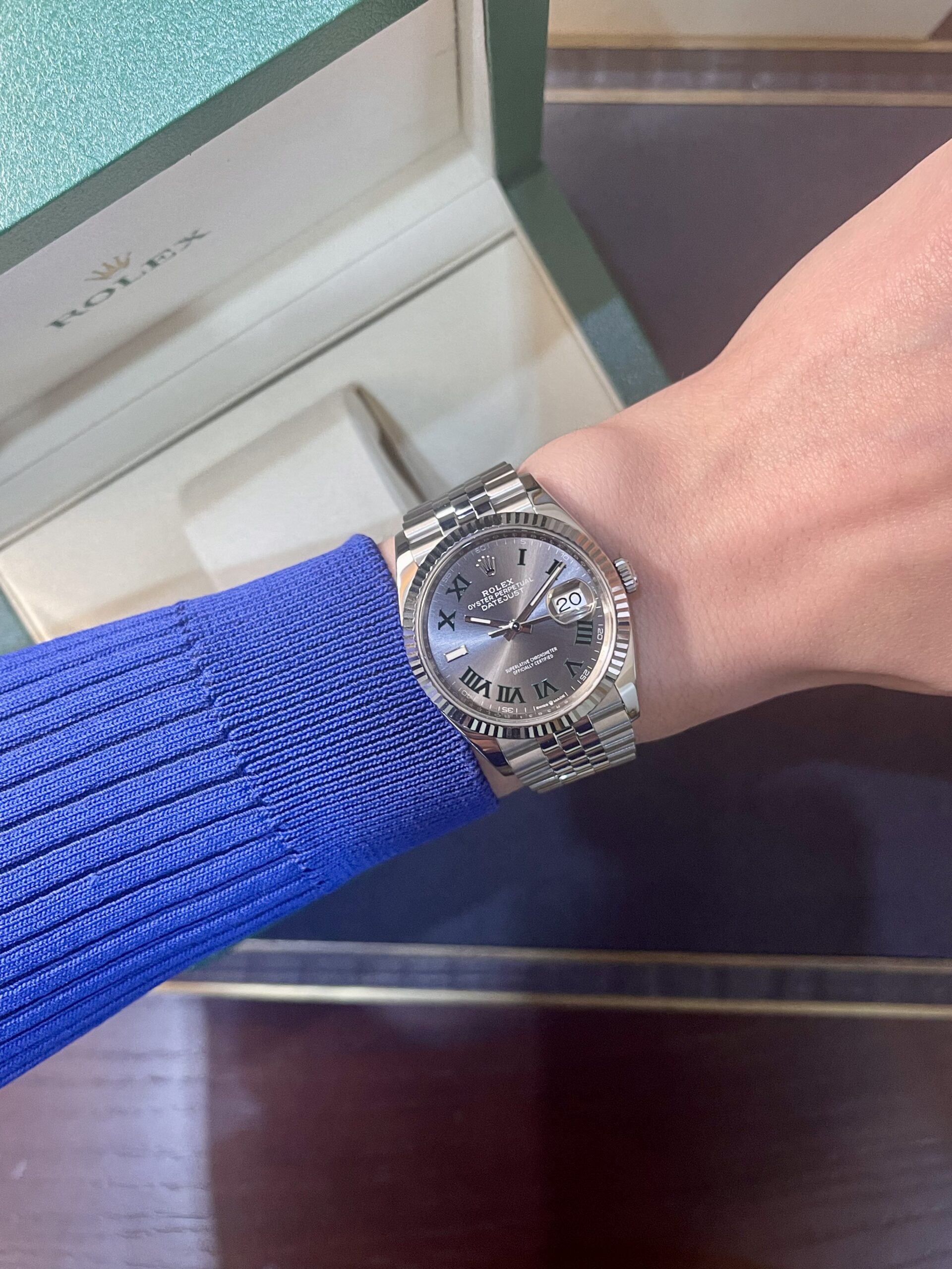 Silver Rolex Datejust on a wrist with a blue sweater sleeve; a Rolex box is visible in the background.