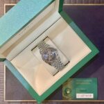 Open green Rolex box reveals a silver Rolex watch with a metal bracelet resting on a beige cushion, date window visible at 3 o'clock.