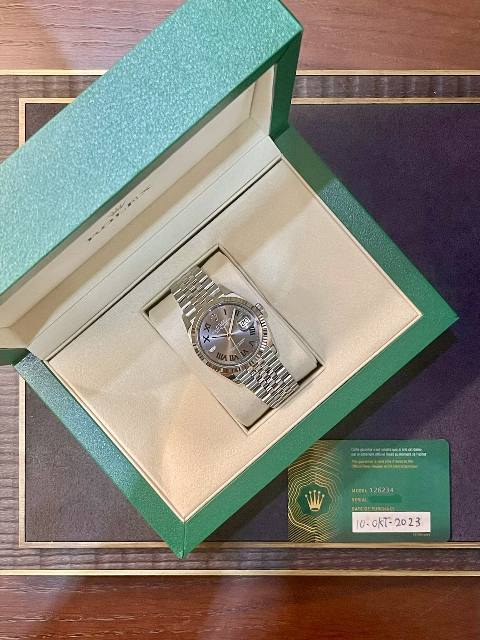 Open green Rolex box reveals a silver Rolex watch with a metal bracelet resting on a beige cushion, date window visible at 3 o'clock.
