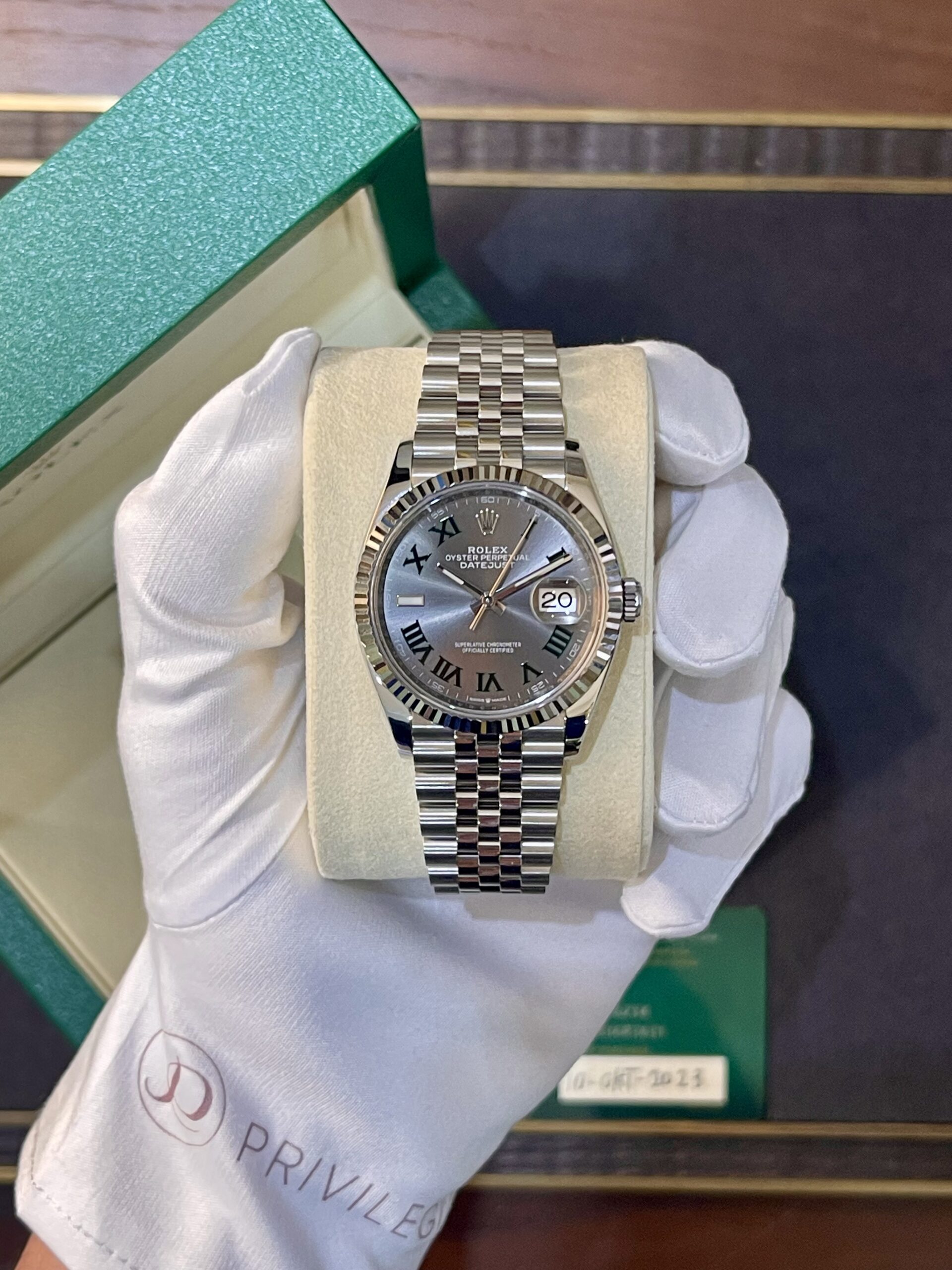 Rolex Datejust stainless steel watch with Roman numeral dial in a green box, held by gloved hand.