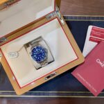 Omega wristwatch in an open wooden display box with cream interior and red trim, blue dial visible.