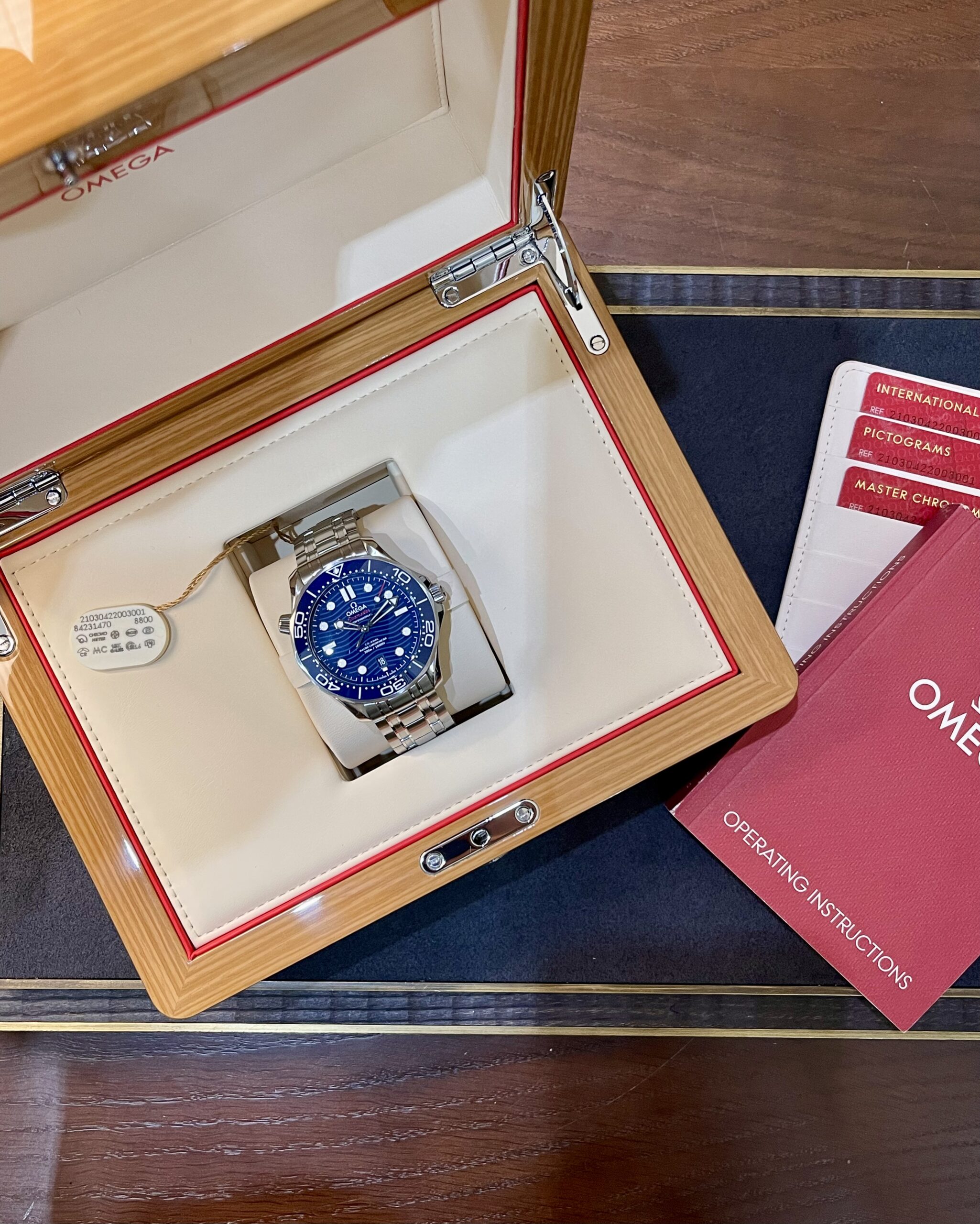 Omega wristwatch in an open wooden display box with cream interior and red trim, blue dial visible.