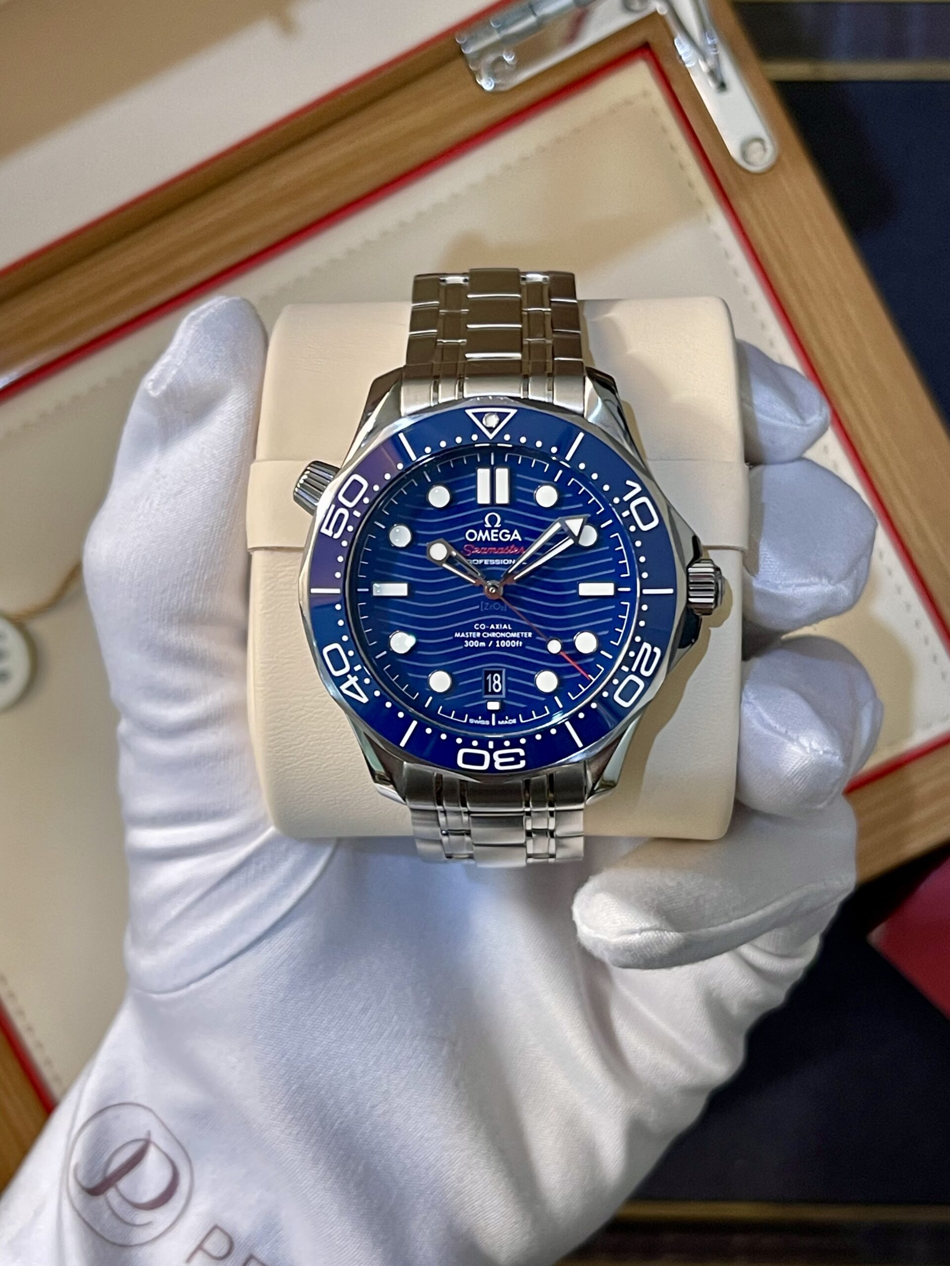 Omega Seamaster diver watch with a blue dial and stainless steel bracelet displayed on a cream cushion in a box.