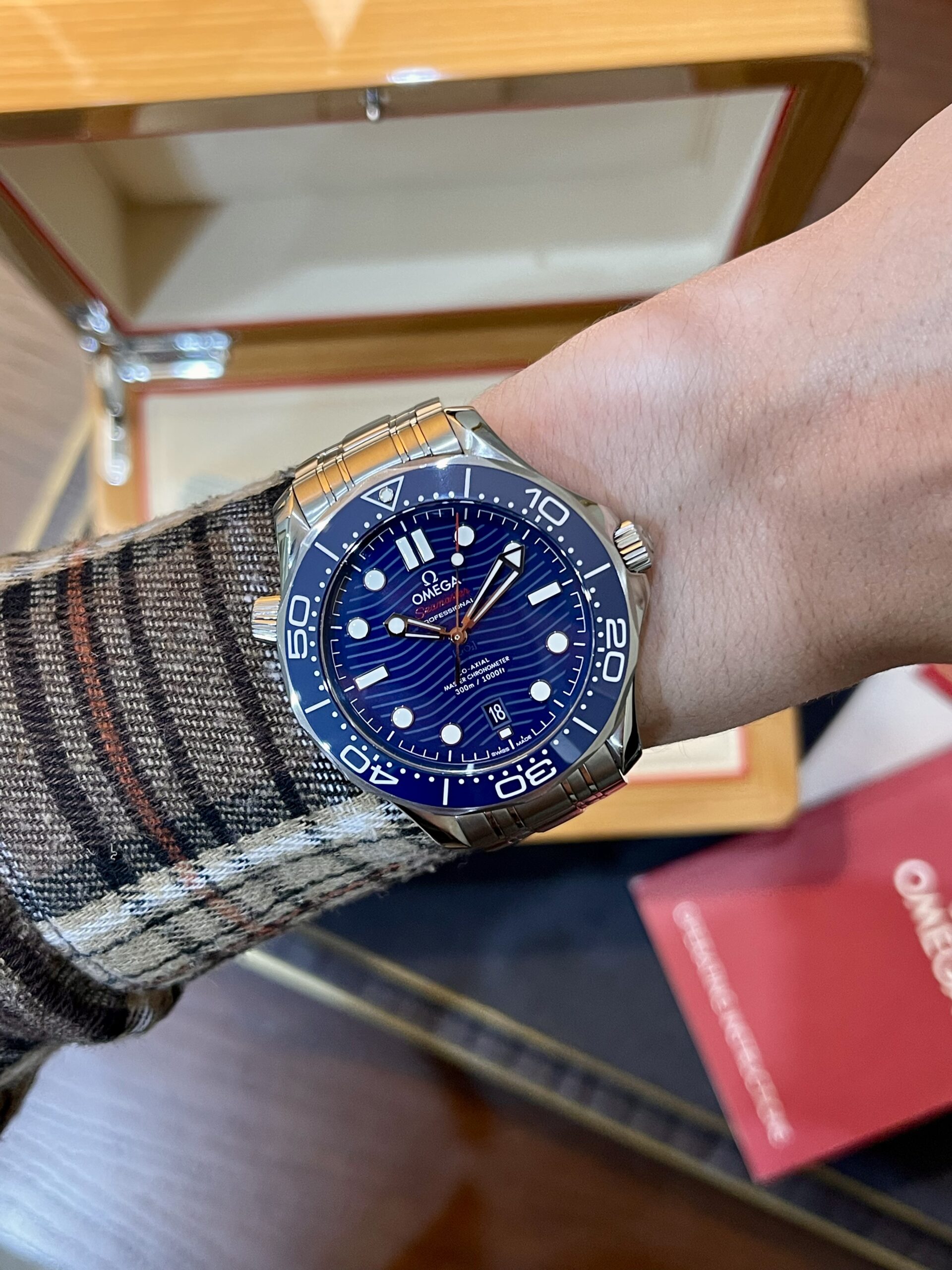 Wrist wearing a blue Omega Seamaster diver's watch with a stainless steel bracelet, plaid sleeve visible in the foreground.